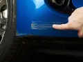 Dito, un dito indica profondi graffi sul parafango blu di un'auto, segno che necessita di riparazione vernice auto.