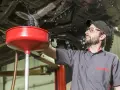 Meccanico con occhiali protettivi e cappellino lavora sotto un'auto sollevata, raccogliendo liquidi in un imbuto rosso. Valutazione pro e contro lavaggio interno motore.
