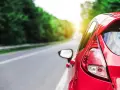 Auto rossa in accelerazione su strada panoramica, con il sole che filtra tra gli alberi.
