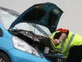 Meccanico con giubbotto ad alta visibilità lavora sul motore di un'auto blu. Nonostante il silenzio delle auto elettriche, il rumore degli attrezzi è forte.
