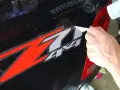 Come togliere colla adesivi: una mano rimuove un adesivo rosso e bianco "Z 4x4" da un'auto nera, usando un phon.