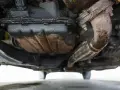 Sotto un'auto, si nota una **perdita olio motore dalla testata**, con accumuli scuri e gocciolamenti visibili sui componenti meccanici e sul tubo di scarico.
