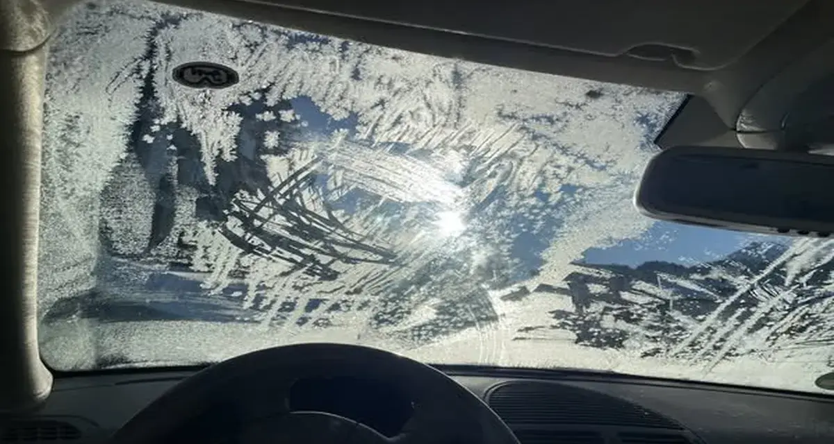 Vetro dell'auto coperto di brina, con segni di tergicristallo. Un sole pallido filtra attraverso il ghiaccio, rendendo difficile vedere. Ecco come sbrinare il vetro della macchina.