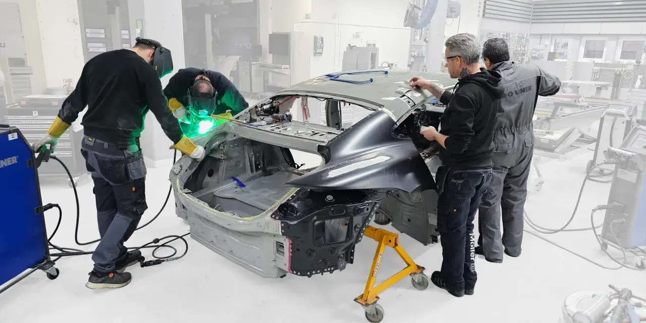 Operai saldano il telaio macchina in officina. Uno di loro usa una saldatrice, mentre gli altri osservano e lavorano sulla carrozzeria.