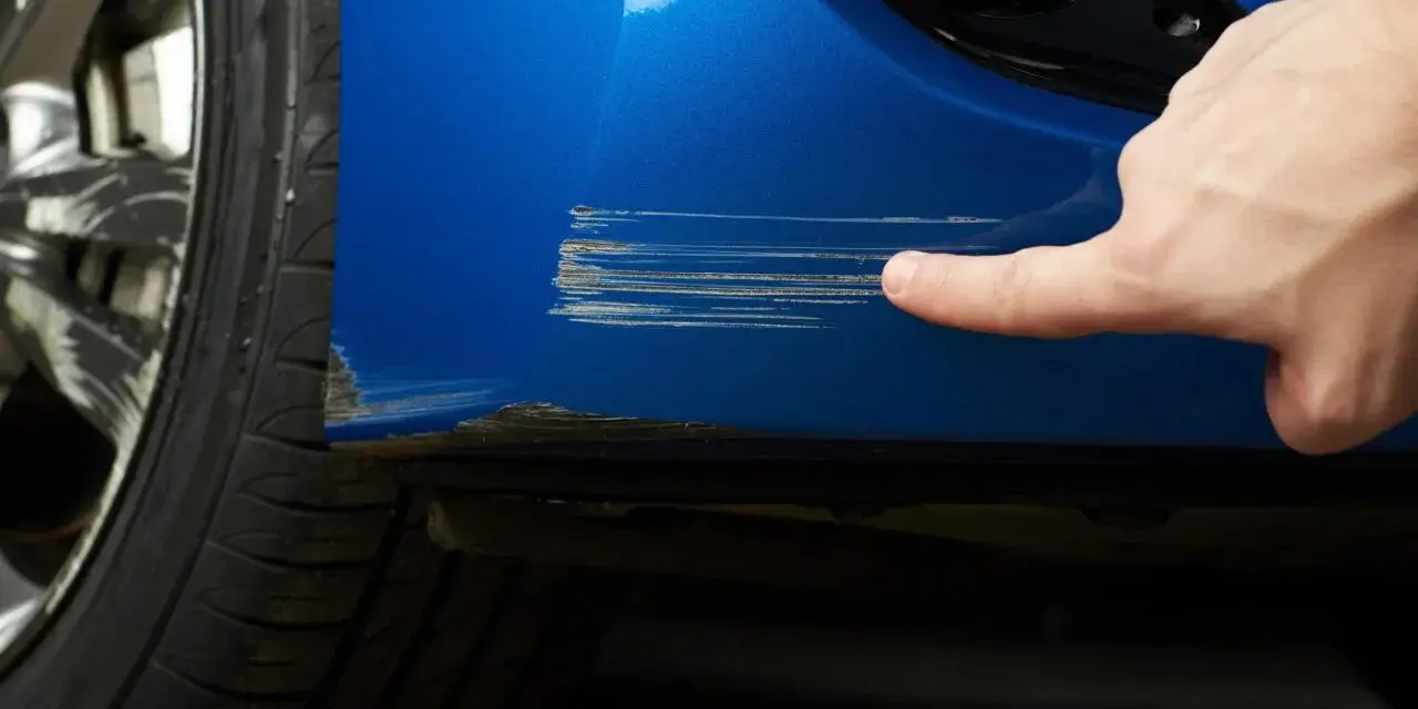 Dito, un dito indica profondi graffi sul parafango blu di un'auto, segno che necessita di riparazione vernice auto.