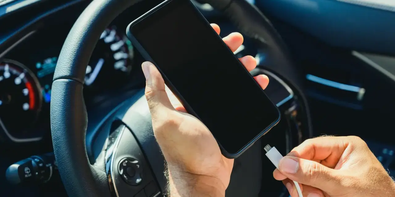 Mano che collega un cavo a uno smartphone, con il volante e il quadro strumenti dell'auto sullo sfondo. Se la batteria è scarica, il quadro si accende.