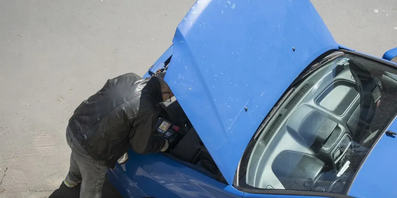 Uomo con giacca in pelle controlla la **macchina ferma** con il cofano aperto, forse un problema alla **batteria**.