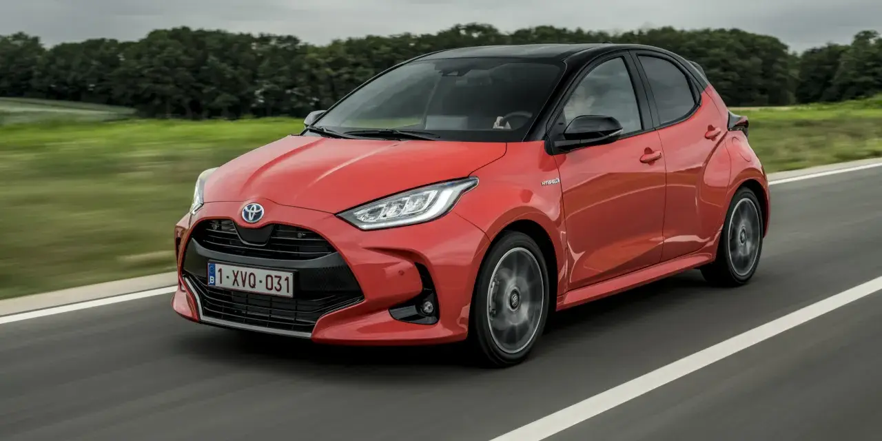 Auto ibrida rossa in movimento su strada, con sfondo sfocato di alberi e cielo nuvoloso. La scritta "Hybrid" è visibile sul fianco.