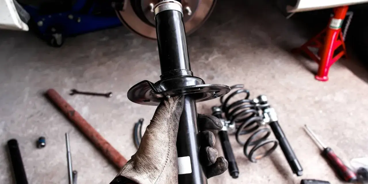 Gloved hand holding a car shock absorber with tools and springs on the ground.