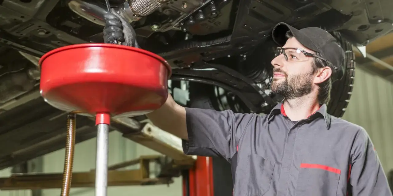 Meccanico con occhiali protettivi e cappellino lavora sotto un'auto sollevata, raccogliendo liquidi in un imbuto rosso. Valutazione pro e contro lavaggio interno motore.
