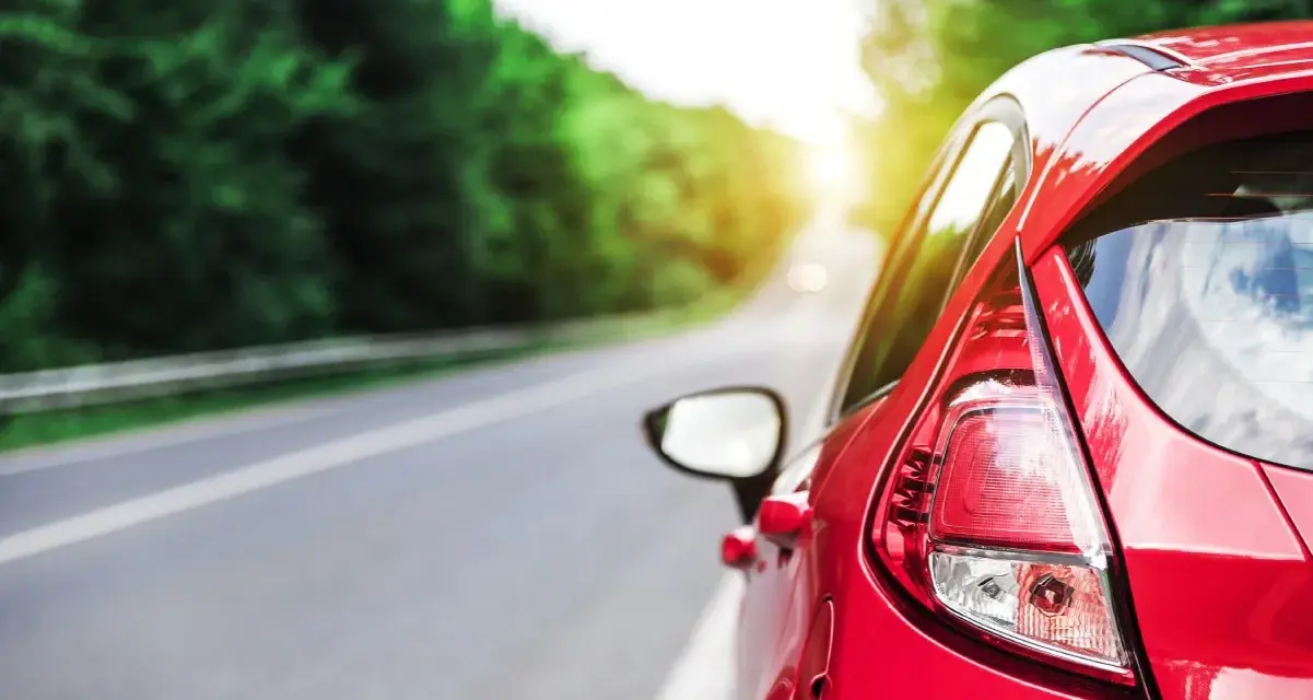 Auto rossa in accelerazione su strada panoramica, con il sole che filtra tra gli alberi.