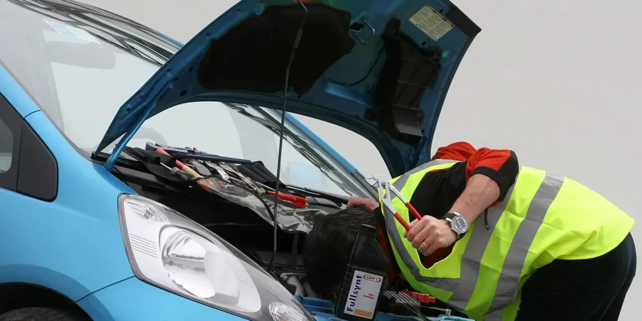 Meccanico con giubbotto ad alta visibilità lavora sul motore di un'auto blu. Nonostante il silenzio delle auto elettriche, il rumore degli attrezzi è forte.