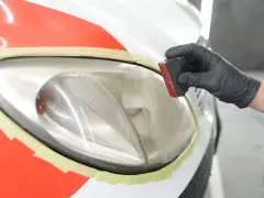Mano con guanto nero lucida un faro opaco dell'auto con una spugna. La carrozzeria è protetta con nastro adesivo.