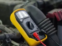 Multimetro giallo collegato per misurare la tensione batteria auto, tra cavi e componenti del motore.