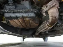 Sotto un'auto, si nota una **perdita olio motore dalla testata**, con accumuli scuri e gocciolamenti visibili sui componenti meccanici e sul tubo di scarico.