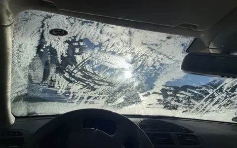 Vetro dell'auto coperto di brina, con segni di tergicristallo. Un sole pallido filtra attraverso il ghiaccio, rendendo difficile vedere. Ecco come sbrinare il vetro della macchina.