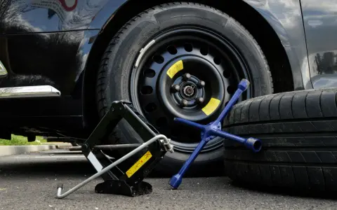 Kit per cambiare una ruota: cric, chiave a stella e ruota di scorta accanto a un'auto nera.