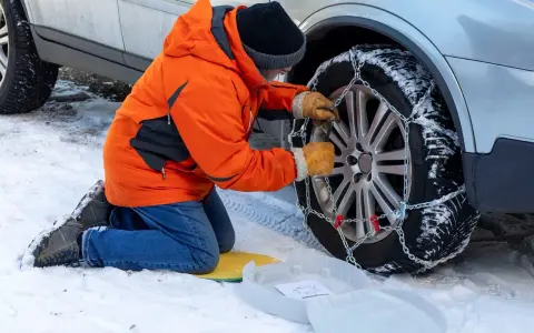 Uomo in giacca arancione monta catene da neve su ruota posteriore. Le catene da neve devono essere montate sulle ruote posteriori per la massima aderenza.