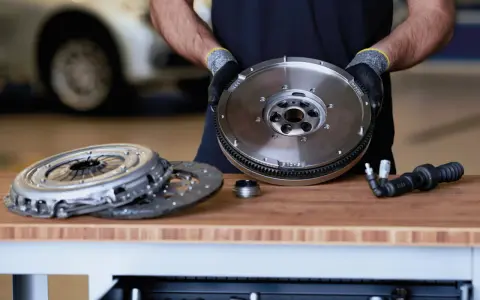 Meccanico mostra un volano auto nuovo, pronto per essere installato. Accanto, un kit frizione e un attrezzo.