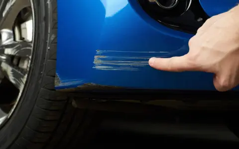 Dito, un dito indica profondi graffi sul parafango blu di un'auto, segno che necessita di riparazione vernice auto.