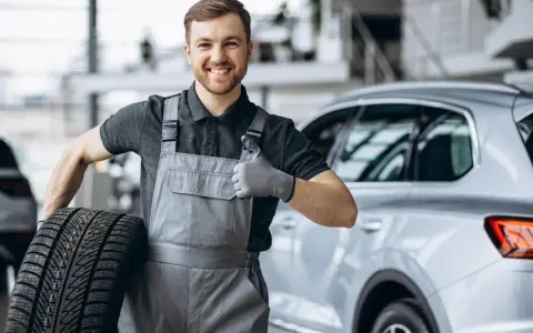 Meccanico sorridente con una gomma primo equipaggiamento in mano, fa il pollice in su accanto a un'auto.