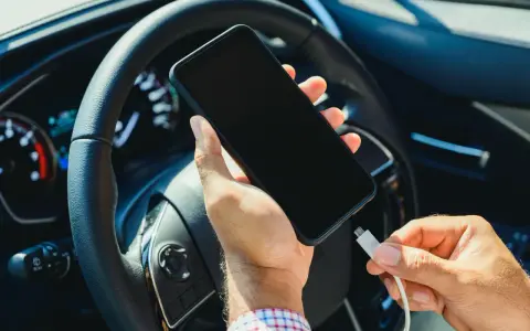 Mano che collega un cavo a uno smartphone, con il volante e il quadro strumenti dell'auto sullo sfondo. Se la batteria è scarica, il quadro si accende.