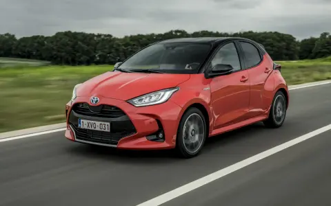 Auto ibrida rossa in movimento su strada, con sfondo sfocato di alberi e cielo nuvoloso. La scritta "Hybrid" è visibile sul fianco.
