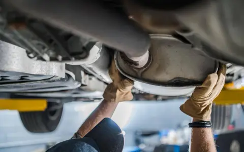Mani guantate lavorano sul sistema di scarico di un'auto, forse sostituendo il catalizzatore auto.