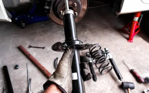 Gloved hand holding a car shock absorber with tools and springs on the ground.