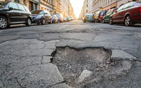 Un cratere nel marciapiede, con auto parcheggiate sullo sfondo. La gomma tagliata dal manto stradale crea un ostacolo pericoloso.