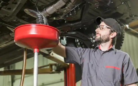 Meccanico con occhiali protettivi e cappellino lavora sotto un'auto sollevata, raccogliendo liquidi in un imbuto rosso. Valutazione pro e contro lavaggio interno motore.