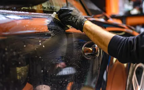 Mano con guanto nero pulisce la carrozzeria lucida di un'auto, rimuovendo la resina con un panno umido e schiumoso.