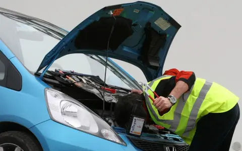 Meccanico con giubbotto ad alta visibilità lavora sul motore di un'auto blu. Nonostante il silenzio delle auto elettriche, il rumore degli attrezzi è forte.