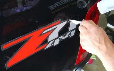 Come togliere colla adesivi: una mano rimuove un adesivo rosso e bianco "Z 4x4" da un'auto nera, usando un phon.