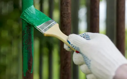 Mano guantata che inizia a verniciare ferro arrugginito con vernice verde brillante, dando nuova vita a una vecchia ringhiera.