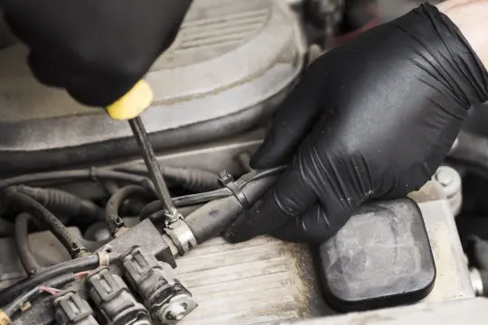 Mani guantate lavorano sul motore a metano, forse per risolvere problemi. Si vede un iniettore e un tubo.