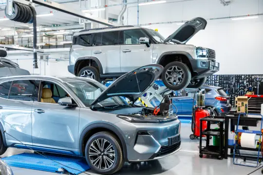 Officina con auto in riparazione. Un SUV grigio è sollevato su un ponte, mentre un'altra auto grigia è a terra con il cofano aperto. Meglio usare poco la macchina si rovina, ma qui la curano.