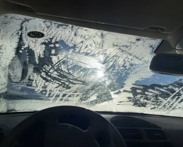 Vetro dell'auto coperto di brina, con segni di tergicristallo. Un sole pallido filtra attraverso il ghiaccio, rendendo difficile vedere. Ecco come sbrinare il vetro della macchina.