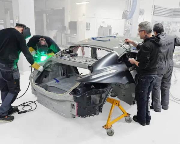 Operai saldano il telaio macchina in officina. Uno di loro usa una saldatrice, mentre gli altri osservano e lavorano sulla carrozzeria.