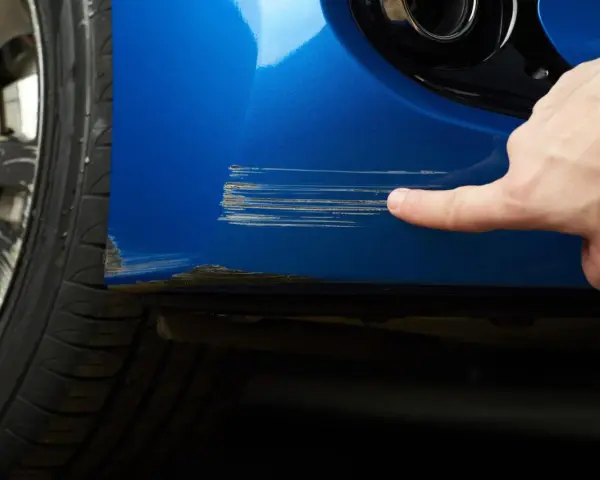 Dito, un dito indica profondi graffi sul parafango blu di un'auto, segno che necessita di riparazione vernice auto.