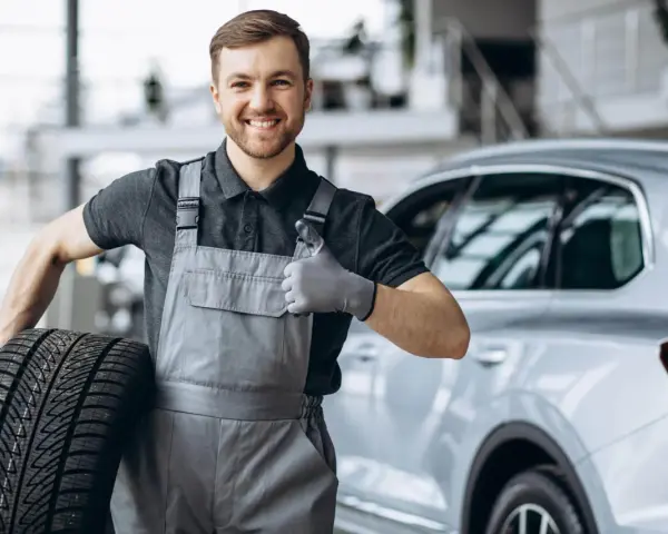 Meccanico sorridente con una gomma primo equipaggiamento in mano, fa il pollice in su accanto a un'auto.