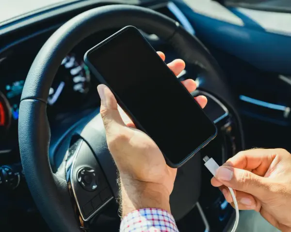 Mano che collega un cavo a uno smartphone, con il volante e il quadro strumenti dell'auto sullo sfondo. Se la batteria è scarica, il quadro si accende.