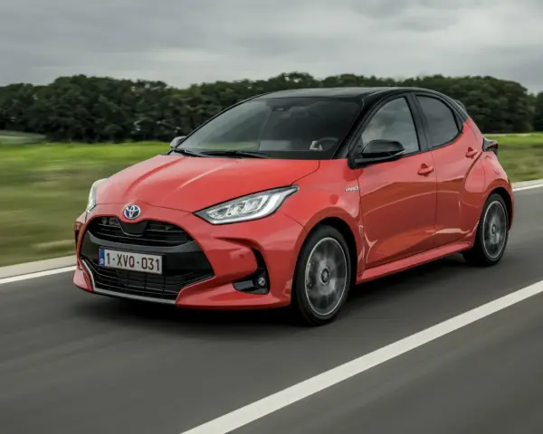 Auto ibrida rossa in movimento su strada, con sfondo sfocato di alberi e cielo nuvoloso. La scritta "Hybrid" è visibile sul fianco.