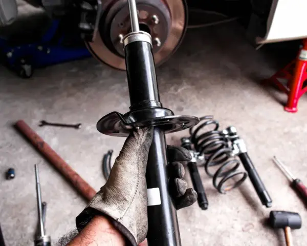 Gloved hand holding a car shock absorber with tools and springs on the ground.