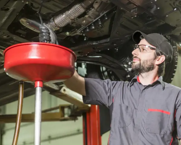Meccanico con occhiali protettivi e cappellino lavora sotto un'auto sollevata, raccogliendo liquidi in un imbuto rosso. Valutazione pro e contro lavaggio interno motore.