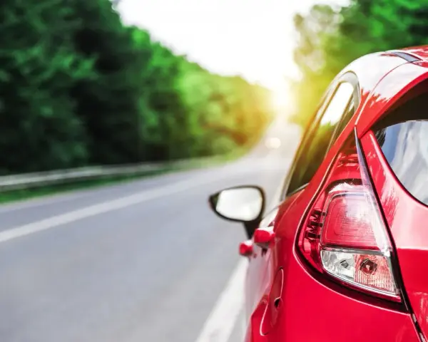 Auto rossa in accelerazione su strada panoramica, con il sole che filtra tra gli alberi.