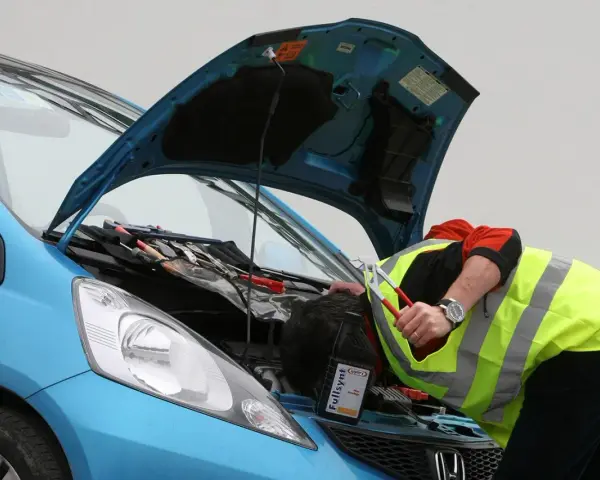 Meccanico con giubbotto ad alta visibilità lavora sul motore di un'auto blu. Nonostante il silenzio delle auto elettriche, il rumore degli attrezzi è forte.