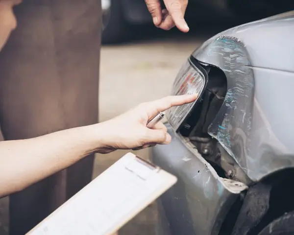 Due mani esaminano i danni alla carrozzeria auto, una indica il faro rotto, l'altra tiene una cartella per la perizia.