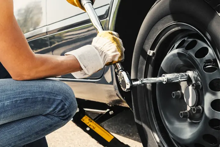 Persona con guanti gialli usa una chiave per serrare i bulloni della ruota di un'auto sollevata da un cric. Il significato di "nm" è la coppia di serraggio.