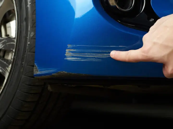 Dito, un dito indica profondi graffi sul parafango blu di un'auto, segno che necessita di riparazione vernice auto.